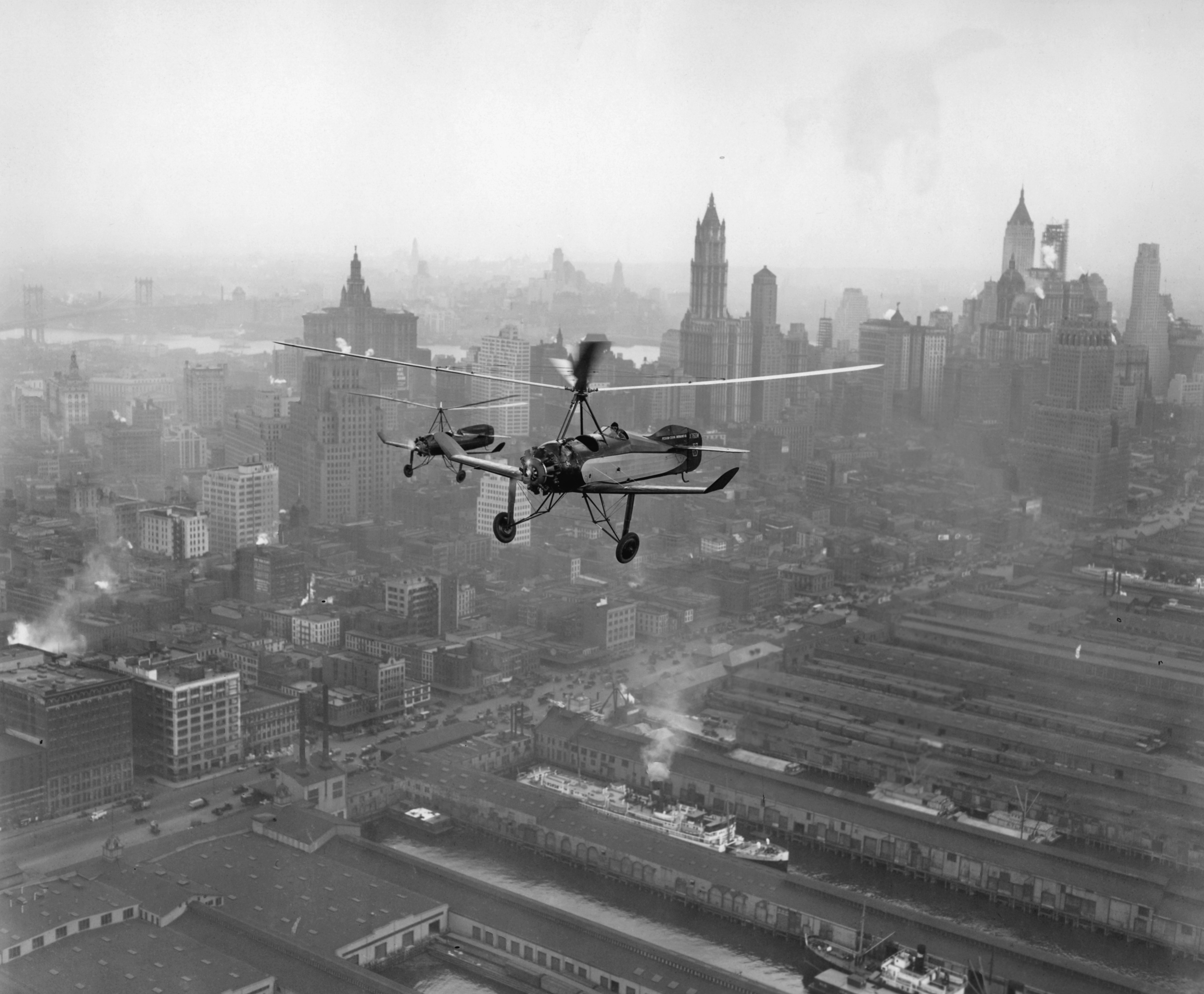 Black and white image of two autogyros flying over a cityscape.