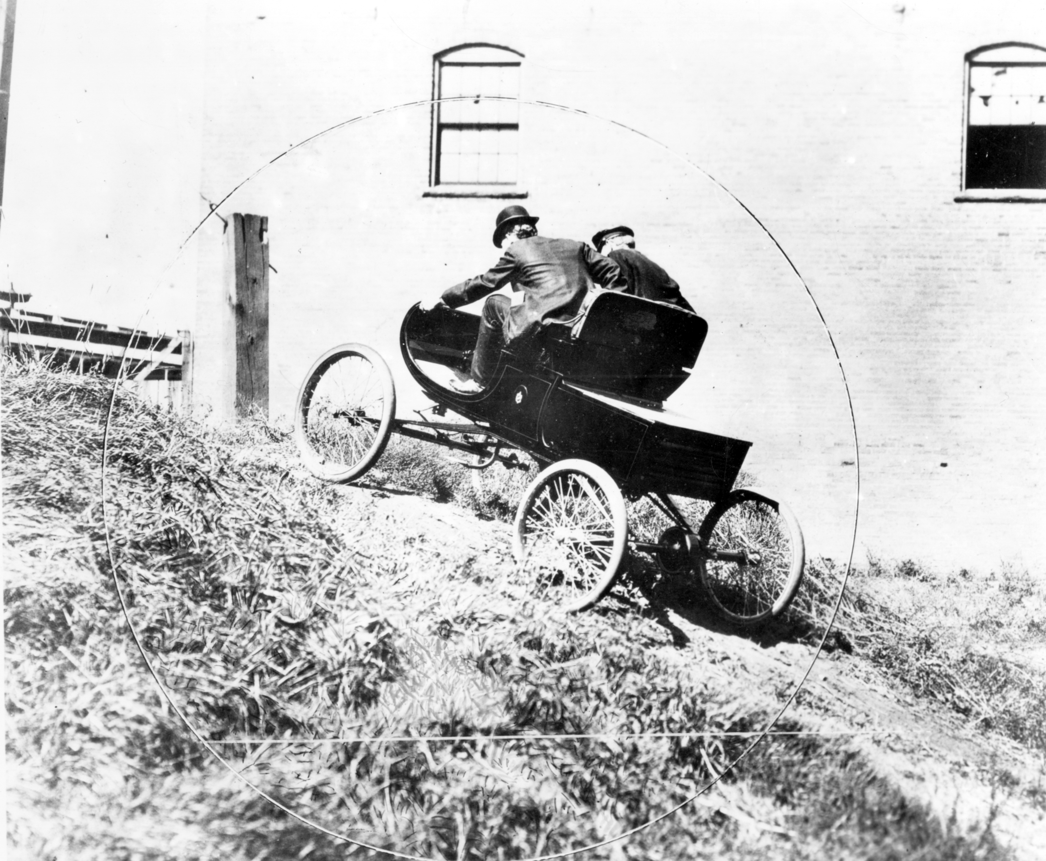 Black and white photograph of two men in a rudimentary automobile driving up a hill.