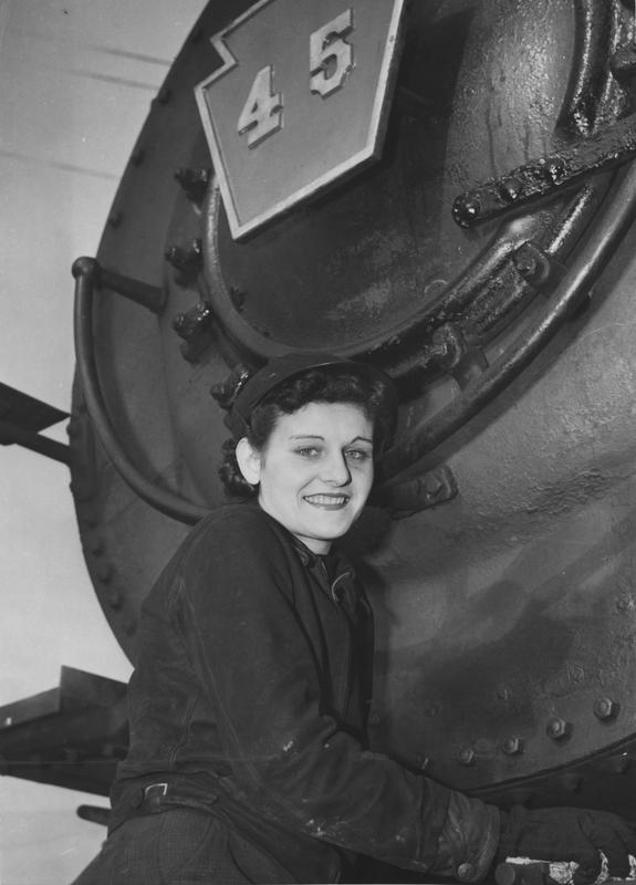 Black and white image of a woman wearing work clothes, posed in front of a locomotive