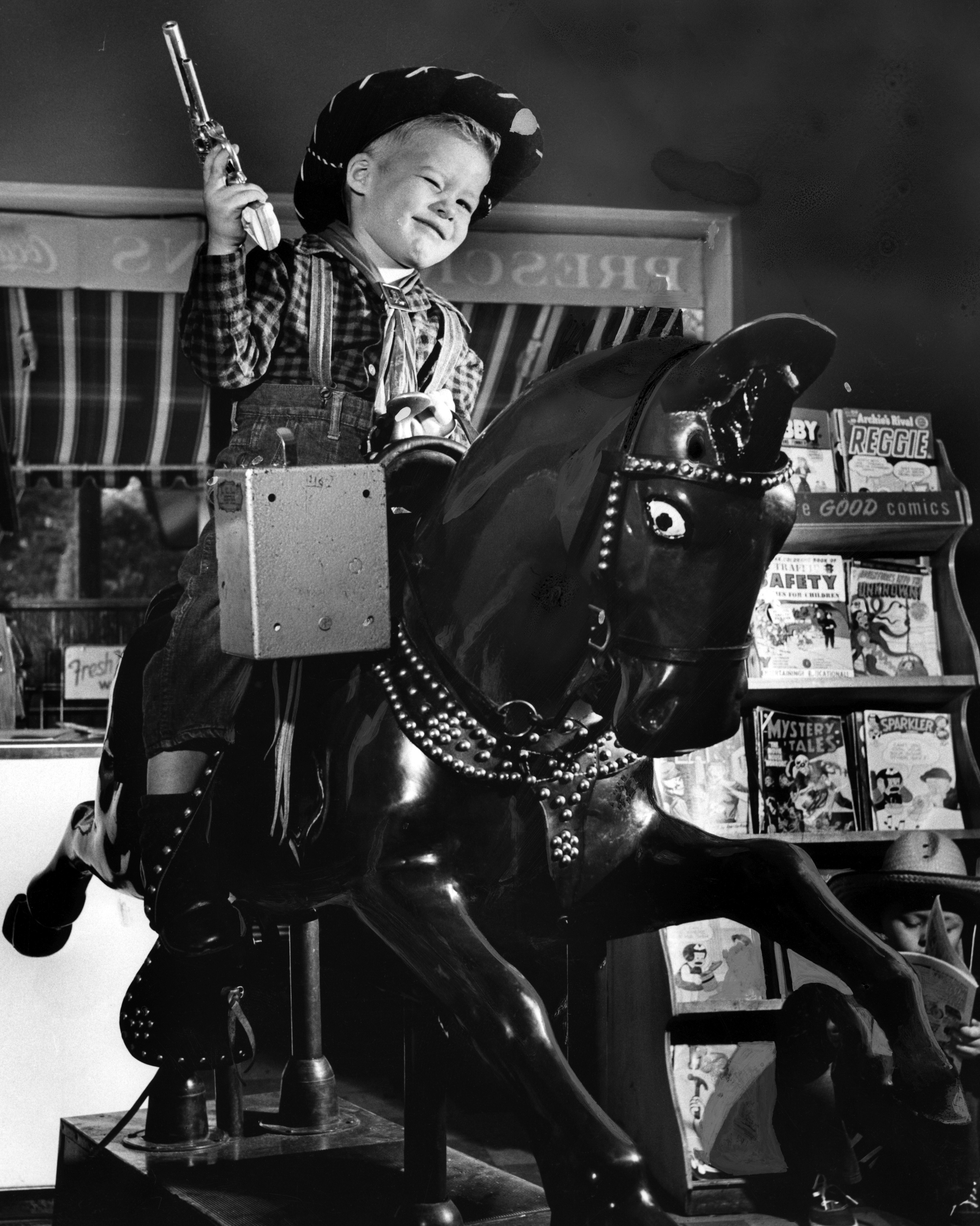 Black and white photograph of a child dressed like a cowboy on a coin-operated horse ride.