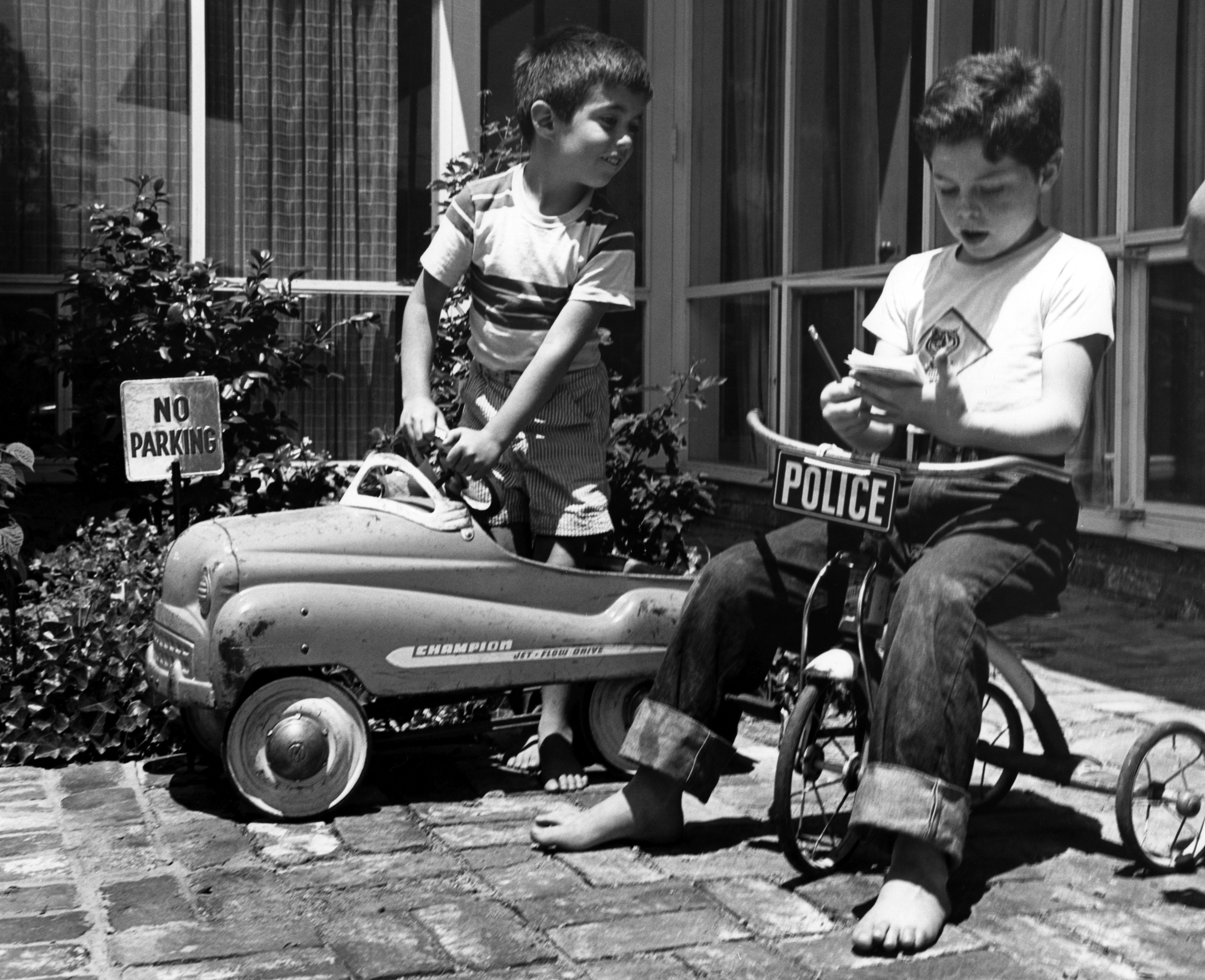 Black and white photograph of two children on a bicycle and a toy car play-acting a traffic stop.