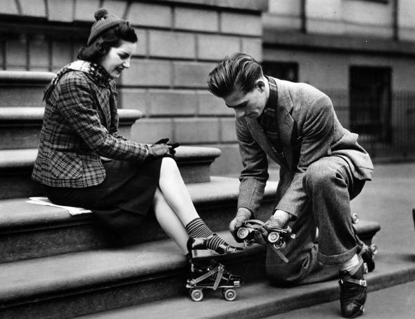 Black and white photograph of a man helping a woman strap roller skates onto her shoes.