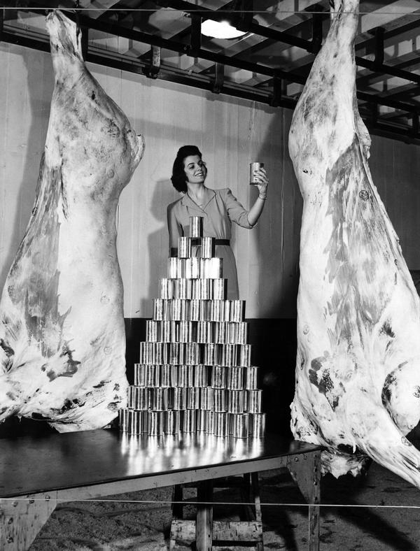 Black and white photograph of a woman with is shown with two sides of beef and a pile of one pound tins that are equal in meat content.