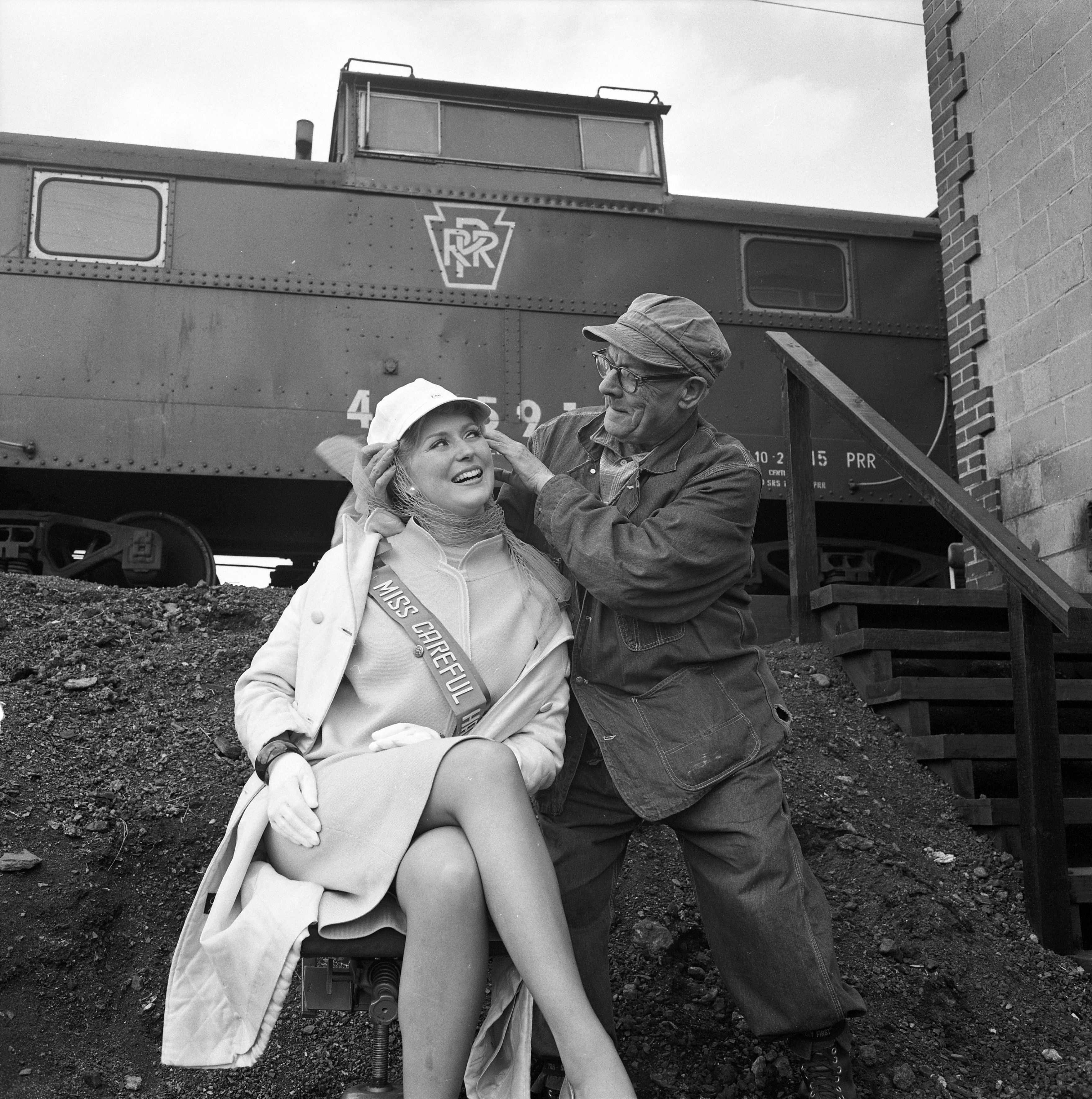 Seated woman in a sash is being crowne with a railroad workers' hat. Train in background.