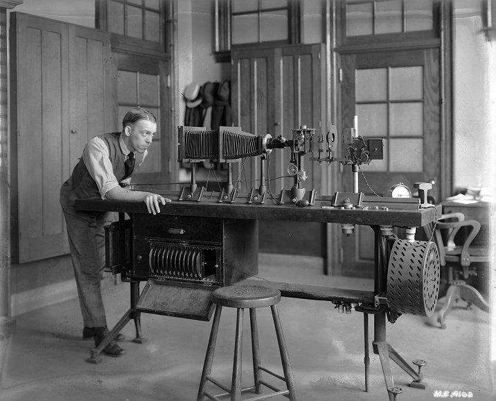 Black and white image of a young man looking into the end of equipment assembled on a tabletop.