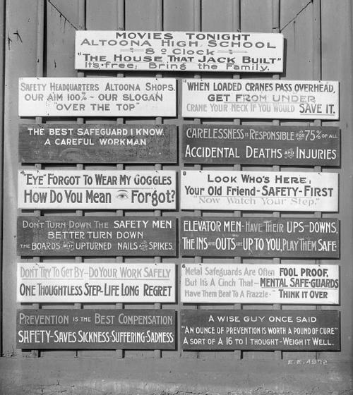 Black and white photo of a collection of signs painted with safety slogans on a wooden wall.