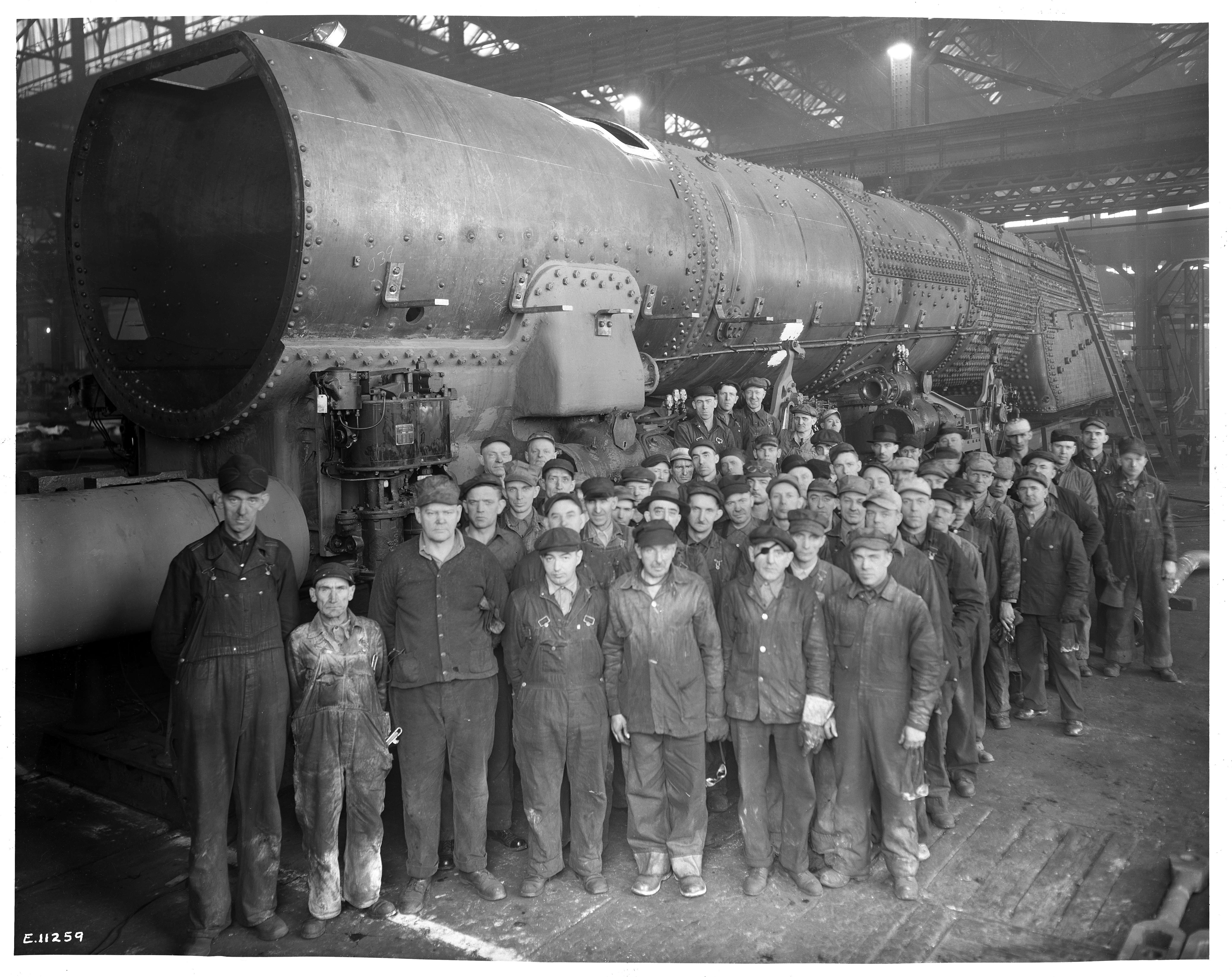 Group photograph of a large number of men in work clothes posed in front of a steam engine boiler