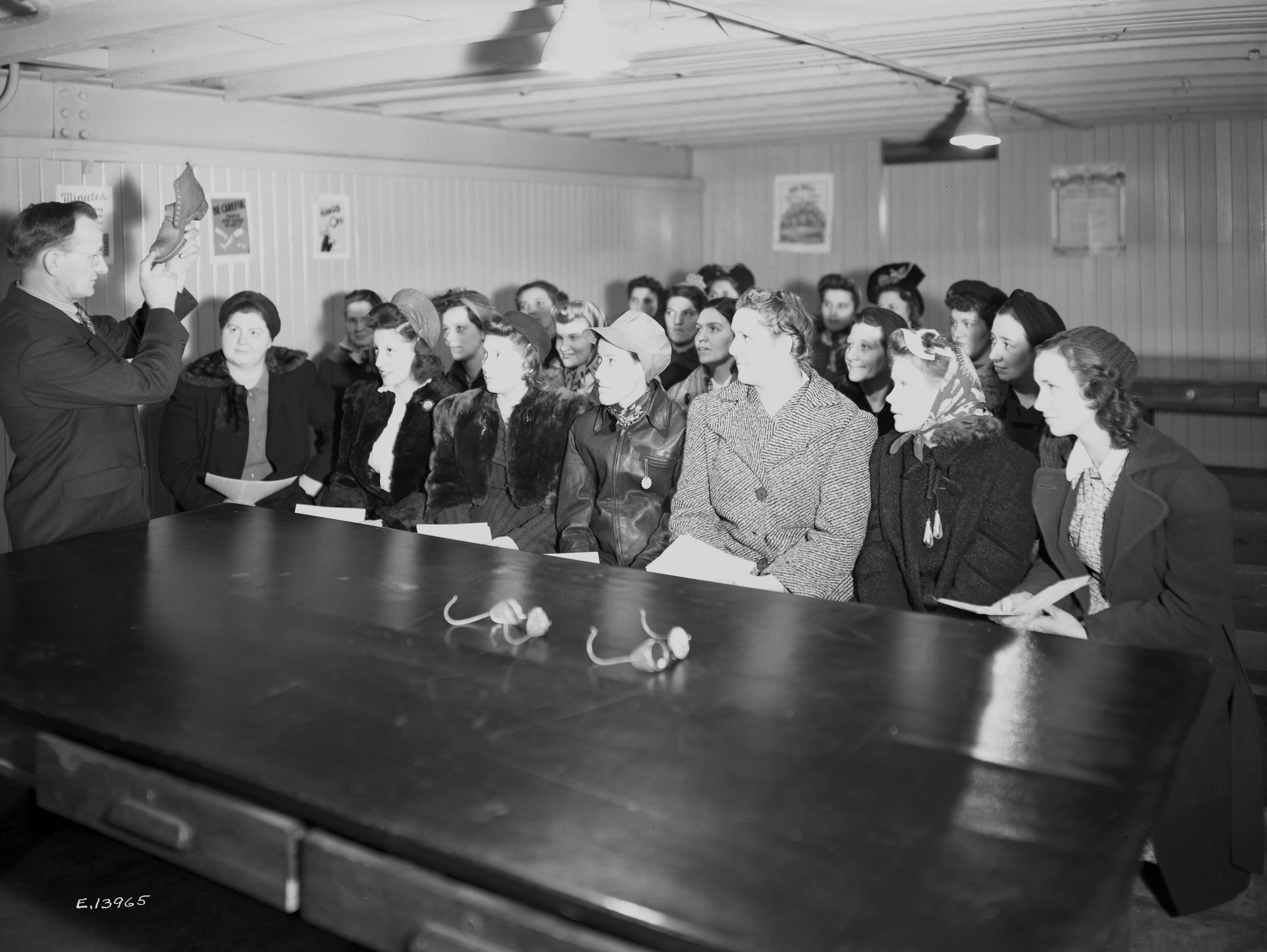 Black and white photograph of a man demonstrating protective goggles and shoes to a classroom of women.