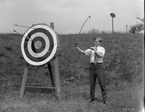 Black and white photograph of a man with a bow and arrow, posed next to a large bullseye target