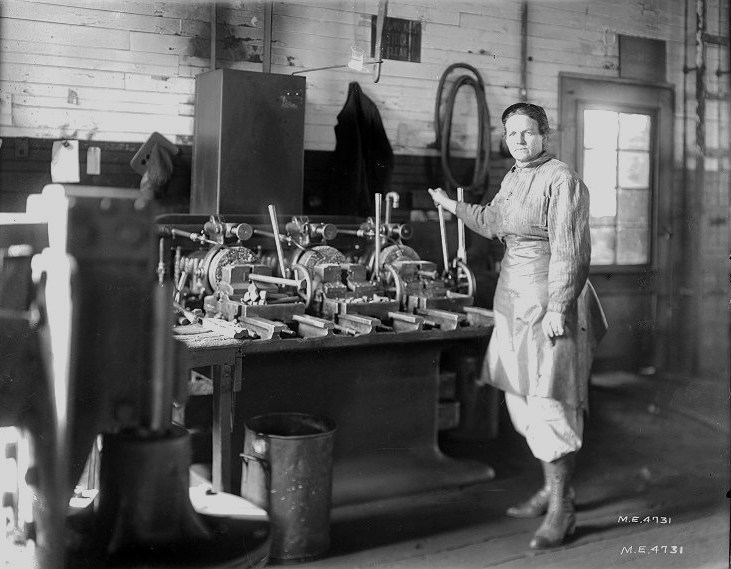 Black and white image of a woman in work clothes standing by a bolt threading machine in a workshop.