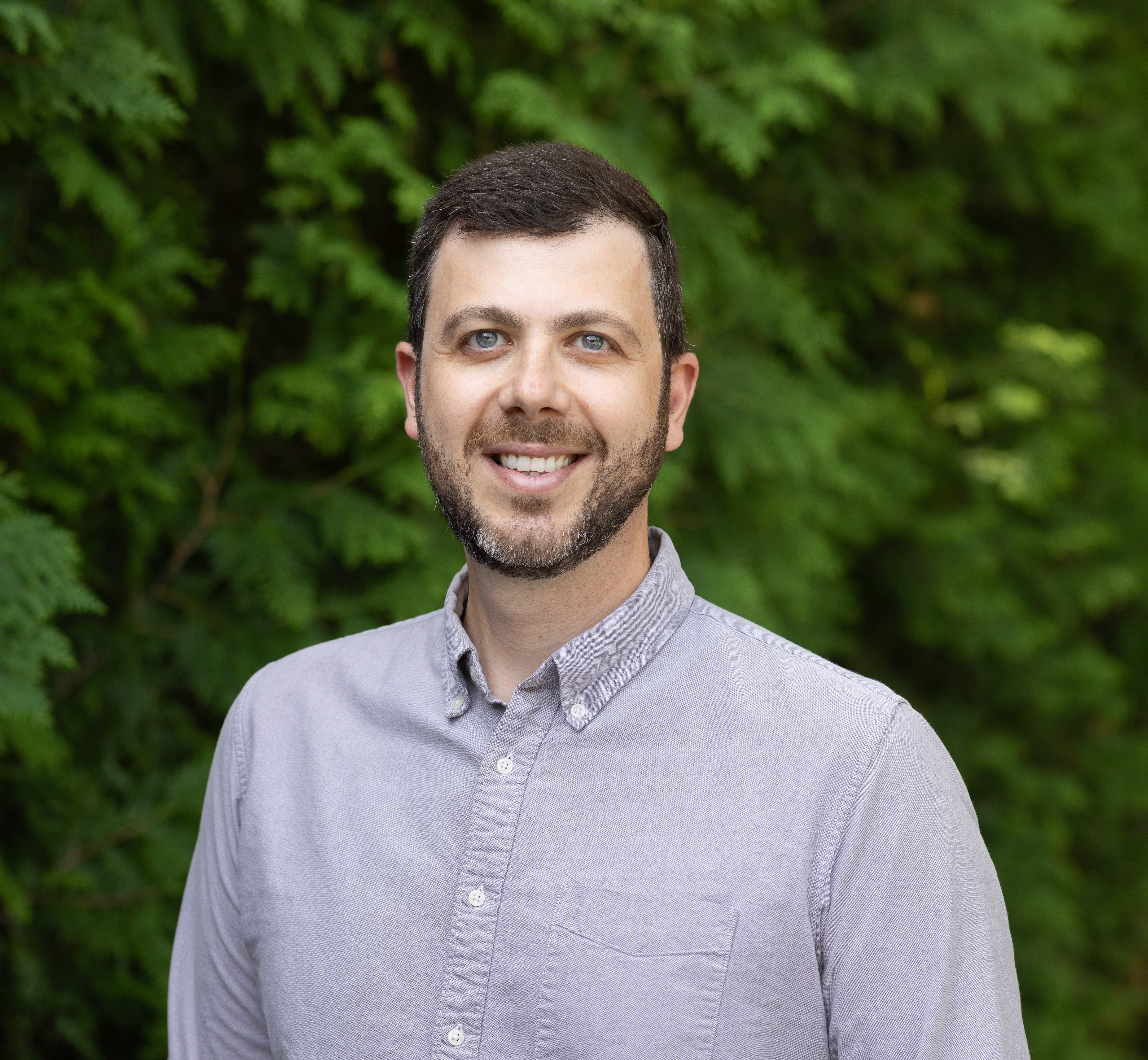 Dylan Gottlieb. A man with brown hair and a beard stand in front of some trees. He is wearing an oxford cloth shirt with a button-down collar.