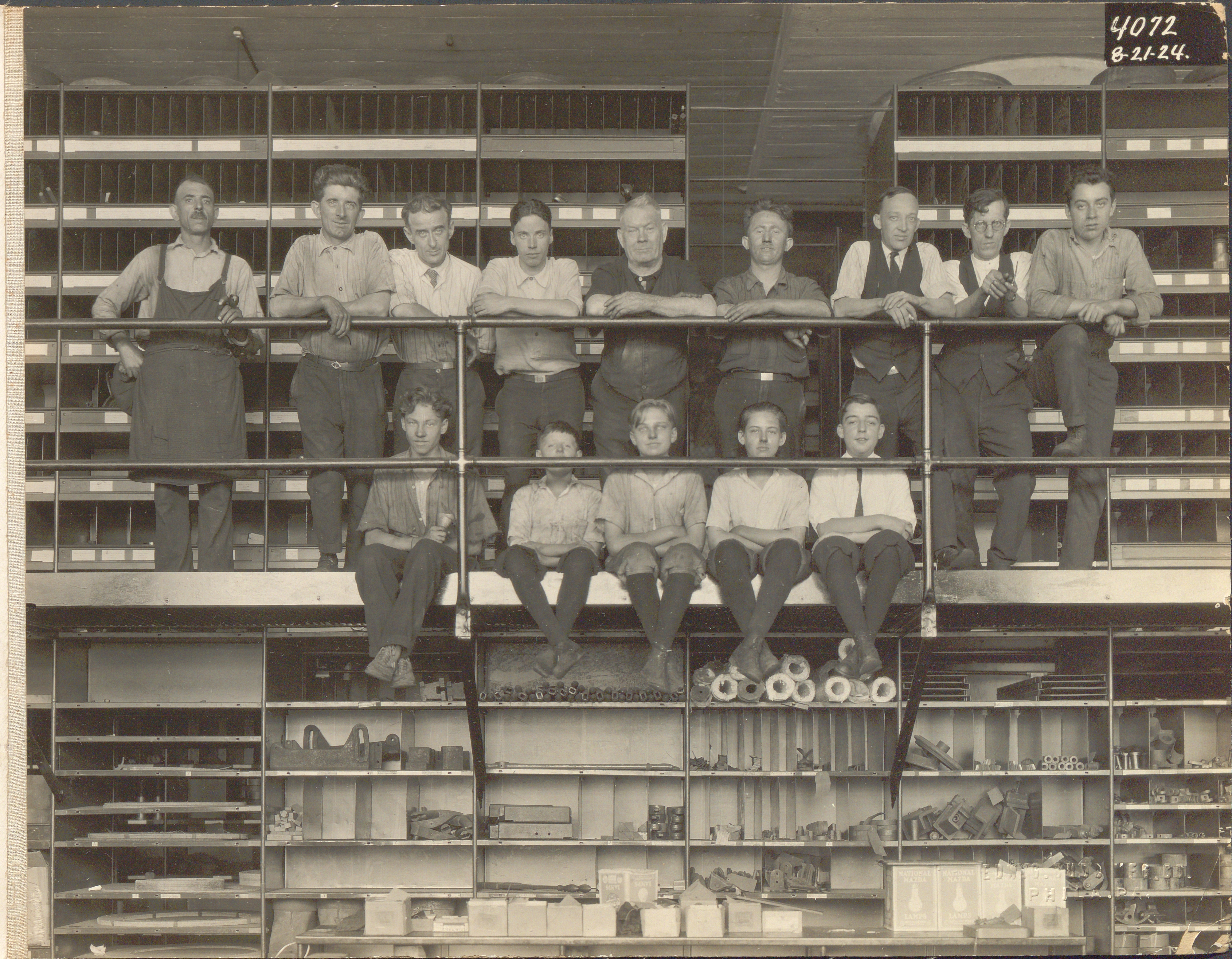 Black and white group photo of men and boys posed on a second-floor walkway of an automobile factory supply area.