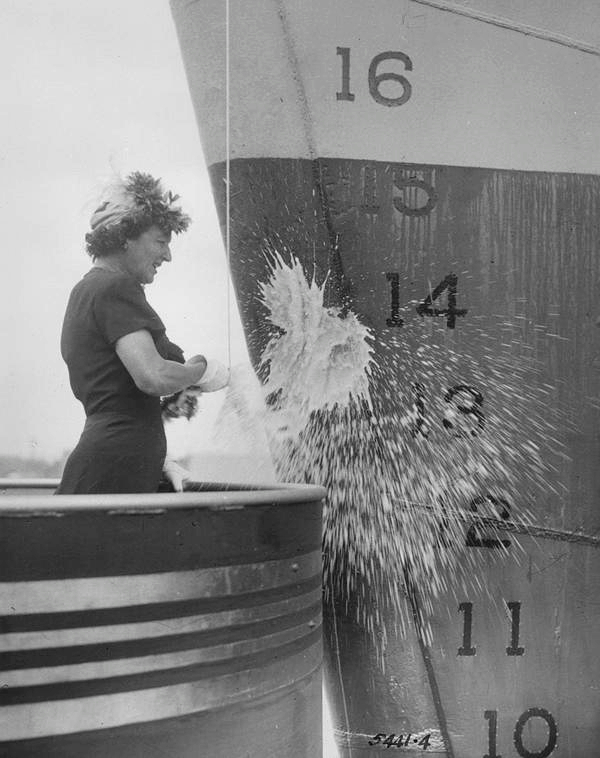 Black and white photograph of a woman christening the hull of a large ship