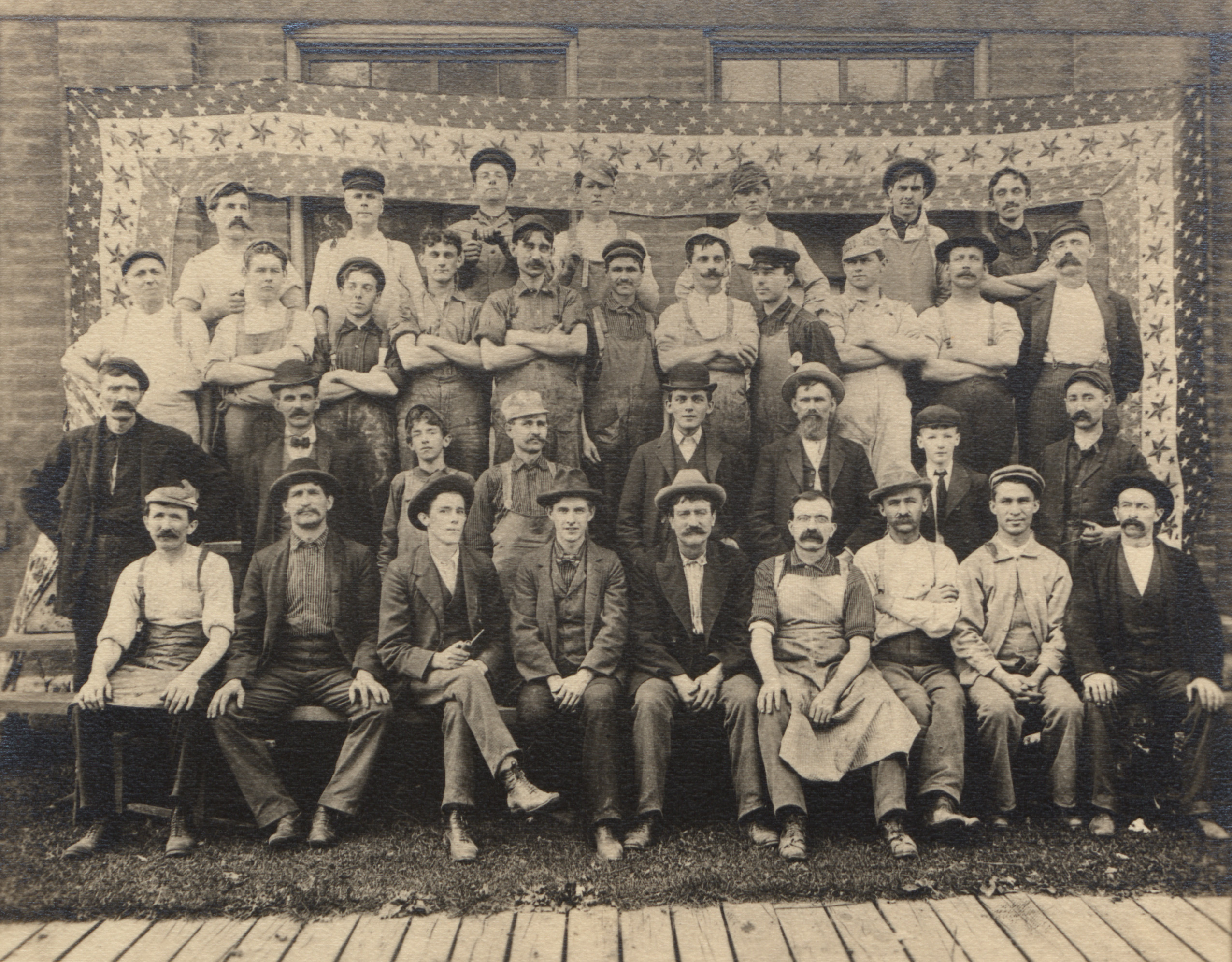 Black and white group portrait of automobile industry workers posed in front of a stars and stripped patterned banner backdrop