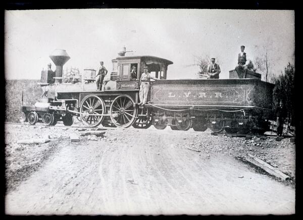 Black and white image of a ca. 1850s wood-burning locomotive, with men posed on and around it.