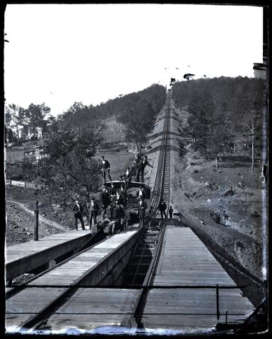 lack and white a image of a railcar at the base of a steep incline, with people aboard and nearby.