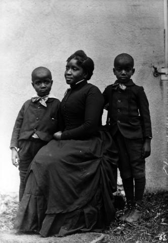 Posed portrait photograph of a seated woman and two standing children, dressed in late 19th-century or early 20th century clothing.