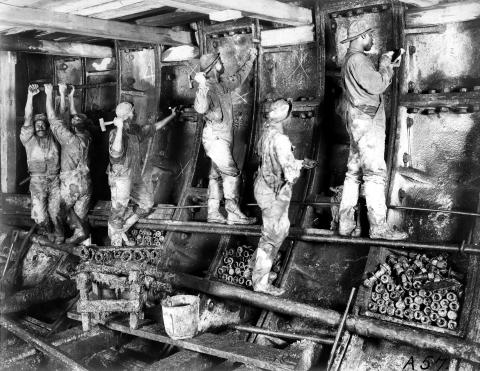 Black and white image of workers hammering and tightening construction elements along the wall of an underground railroad tunnel.