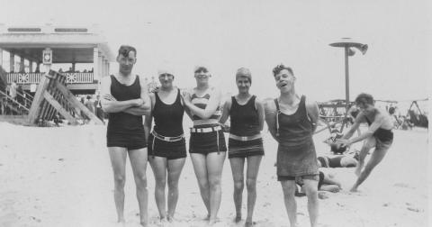 A black and white photograph depicts a group of men and women on the beach in 1920s swimwear