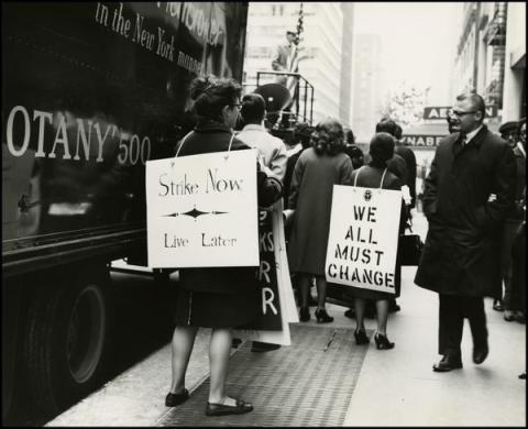 Street scene of a protest with women wearing signs reading "Strike Now - Live Later" and "We all must change."