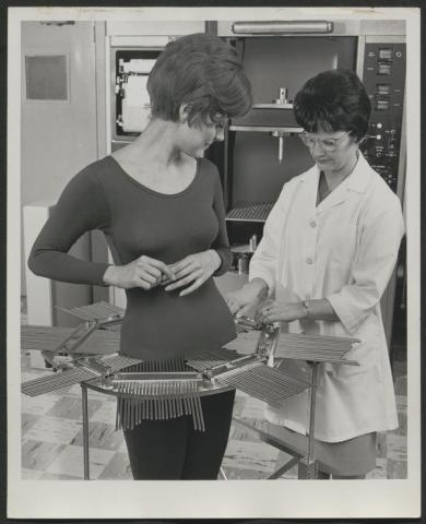 Black and white photograph of a woman in a laboratory measuring another woman's hip area with a contour meter apparatus.