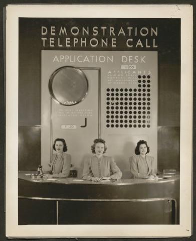 Black and white image of three women at a demonstration telephone call application desk.
