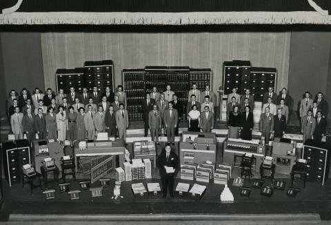 Black and white photo of a large group of men and women with office equipment and paperwork.