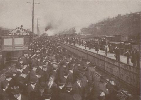 Photograph of a large group of men in hats waiting at a train station in East Pittsburg