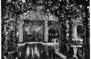 Black and white image of an interior pool surrounded by seating and covered by flowered pergolas