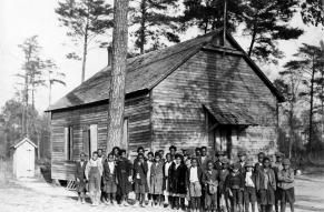 Black and white photo of young, Black schoolchildren outside a one room schoolhouse.