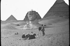 Black and white image of a photographer capturing a person sitting atop the at Sphinx, with the Pyramid of Chephren in the background