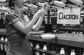 Black and white image of a woman working in front of an array of spools being filled with Dacron fiber. A sign nearby reads 'Dacron'