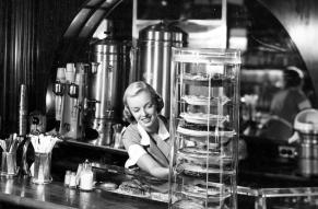 Black and white photograph of a woman at a diner adjusting a display case for pies.