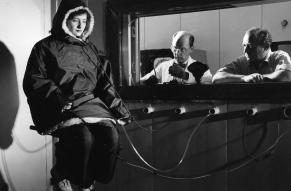 Black and white image of a woman in a large parka seated in a laboratory while two researchers observe behind a viewing window.