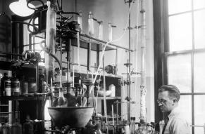 Black and white image of man standing in front of a large array of chemical laboratory apparatus.