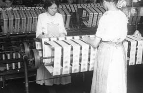 Black and white image of two young women working at a stand of silk manufacturing machinery.