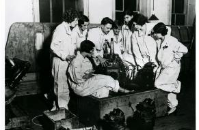 Group of women in flights suits gathered around two men demonstrating the parts of an aircraft motor.