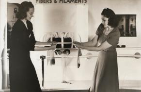 Black and white image of two women at an exhibition booth pulling on either end of a leg of nylon stocking.