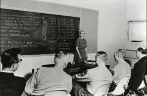 Black and white image of Grace Hopper presenting on COBOL in front of blackboard