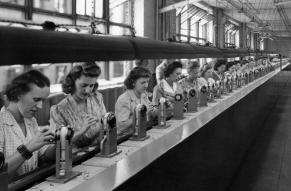 Photograph of a long line of women in a factory, each working on a machine part in front of them on a conveyor belt.