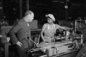 Black and white photograph of a man speaking to a woman operating a lathe.