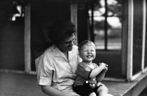 Black and white image of a smiling toddler seated on a woman's lap on a porch