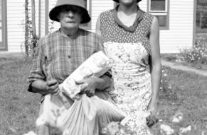 Black and white photo. Two people posed behind a waist-high flowering plant. Home and yard in the background.