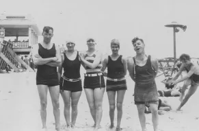 A black and white photograph depicts a group of men and women on the beach in 1920s swimwear