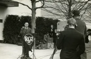 A black and white photograph of three men, one standing behind an RCA podium and the other two filming the first man speaking.