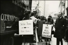 Street scene of a protest with women wearing signs reading "Strike Now - Live Later" and "We all must change."