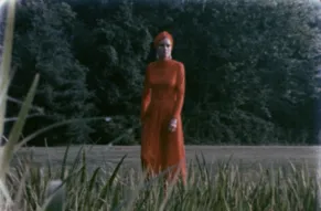 A woman dress in a red dress standing in a field and looking at the camera.