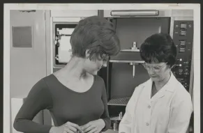 Black and white photograph of a woman in a laboratory measuring another woman's hip area with a contour meter apparatus.