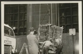Black and white photograph of men loading a large computer component onto a hoist for a crane.