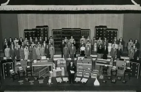 Black and white photo of a large group of men and women with office equipment and paperwork.
